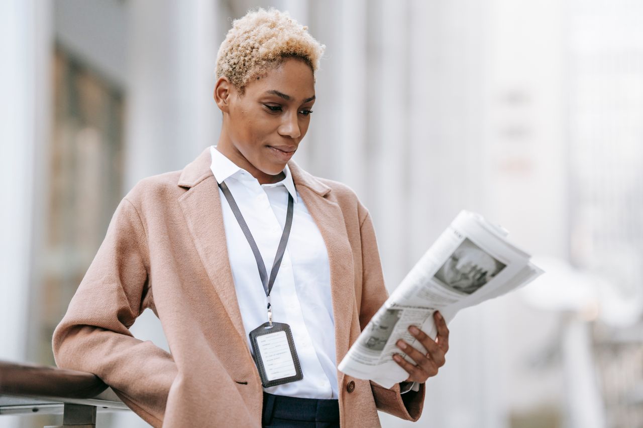 Serious young African American female entrepreneur with badge in elegant brown coat standing near railing and reading newspaper