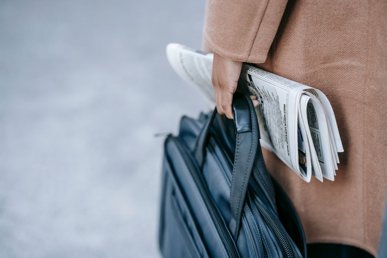 heros-img Crop anonymous female entrepreneur in elegant brown coat with laptop in bag and newspaper